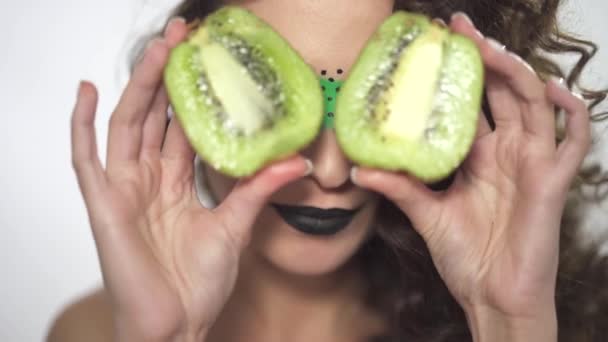 Портрет молодой красивой кудрявой девушки с творческим макияжем, покрывающим глаза двумя фруктовыми частями киви — стоковое видео