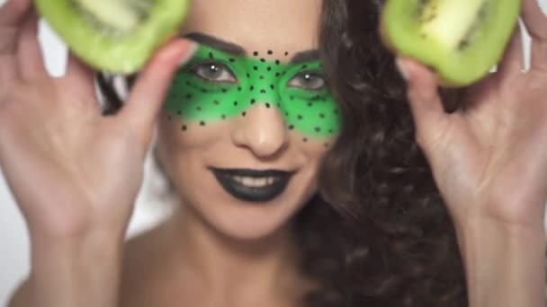 Retrato de la joven hermosa chica rizada con creativo maquillaje cubriendo los ojos con dos partes afrutadas de kiwi — Vídeos de Stock