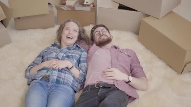 陽気な若者は、ふわふわの絨毯に横になっているアパートのインテリア レイアウトをについて説明します。結婚されていたカップルは、新しい家に移動します。近くの多くのボックスを床に横たわって若いカップルを見てください。. — ストック動画