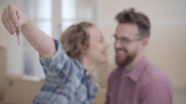 Portrait of a couple in love, they just bought a house and moving. Woman extends a hand with keys to the camera, man kissing her in cheek. Focus changes from keys to people faces — Stock Video