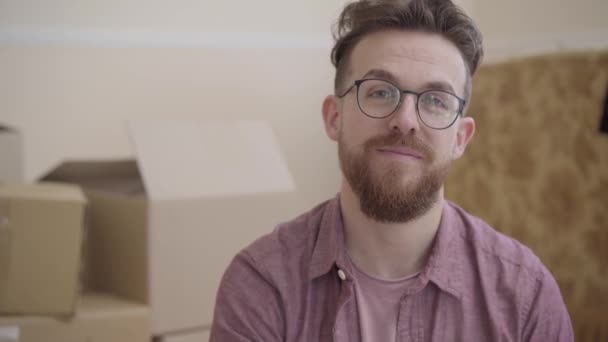 Retrato de homem barbudo em óculos demonstrando chave de casa olhando na câmera com largo sorriso. O homem positivo muda-se para uma nova casa. As caixas estão em segundo plano — Vídeo de Stock