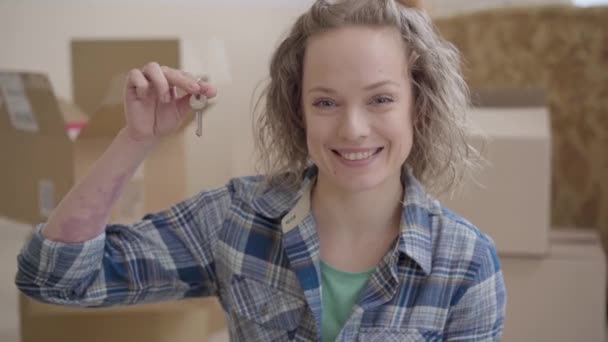Retrato de mujer linda en camisa a cuadros que muestra la llave de la casa mirando en la cámara con amplia sonrisa. La mujer positiva se muda a un nuevo hogar. Las cajas están en el fondo — Vídeo de stock