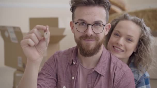 Portrait of a young family moved to a new home. Bearded man with glasses showing the keys to a new home. Wife hugging her husband sitting joyful next. Concept of happy family — Stock Video
