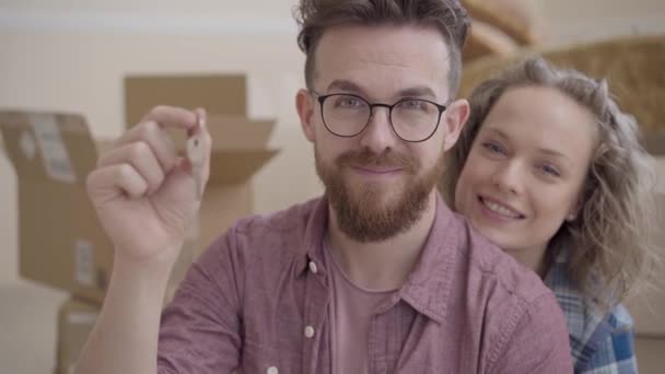 Retrato de una familia joven se mudó a un nuevo hogar. Hombre barbudo con gafas que muestran las llaves de un nuevo hogar. Esposa abrazando a su marido sentado alegre siguiente. Concepto de familia feliz — Vídeo de stock