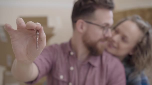 Portrait of a couple in love, they just bought a house and moving. Bearded man in glasses shows the key, looking at his wife then in camera, smiling. Cute woman hugs him. Focus changes — Stock Video