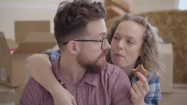 Retrato de una mujer guapa come galletas, abrazándose por detrás del hombre barbudo con gafas de cerca. Cajas desempaquetadas están en el fondo. La pareja casada se muda a un nuevo hogar. Descanso familiar juntos — Vídeo de stock