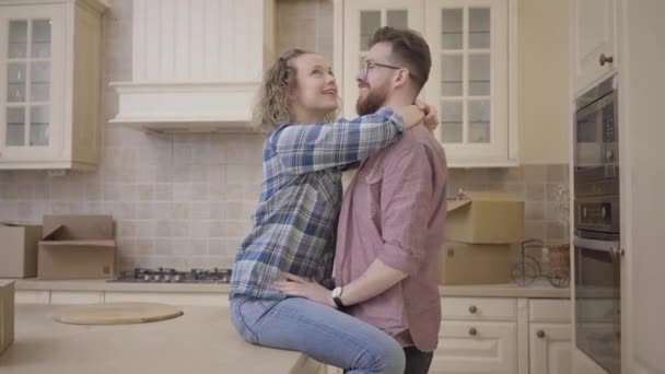 Homem barbudo fica perto de mulher bonita sentada na mesa na grande cozinha meio vazia. A mulher abraça o marido, beijam-se. Casal casal se muda para uma nova casa — Vídeo de Stock