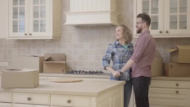 Молодая счастливая семья целуется стоя на кухне в своей новой квартире. Бородатый муж в очках в клетчатой рубашке и его жена с кудрявыми светлыми волосами в джинсах и клетчатой рубашке. Концепция счастья — стоковое видео