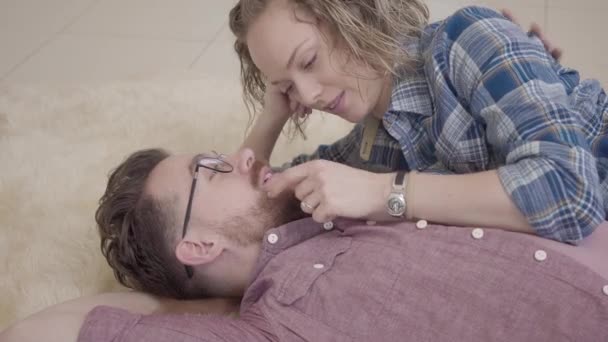 Primer plano retrato de hombre y mujer acostados juntos en una alfombra esponjosa y hablando. La señora toca la barba y el pecho del marido. Barbudo hombre en gafas y bonita mujer con pelo rizado pasar tiempo juntos — Vídeos de Stock