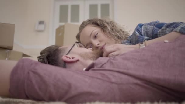 Close up portrait of man and woman lying on the floor and talking in his new apartment. Bearded man in glasses and pretty woman with curly hair spend time together — Stock Video