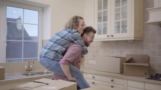 Bearded man carries pretty woman sitting on his back in the kitchen. Woman hugs husband and shows him direction with her finger. Married couple have a fun at new home, unpacked boxes everywhere — Stock Video