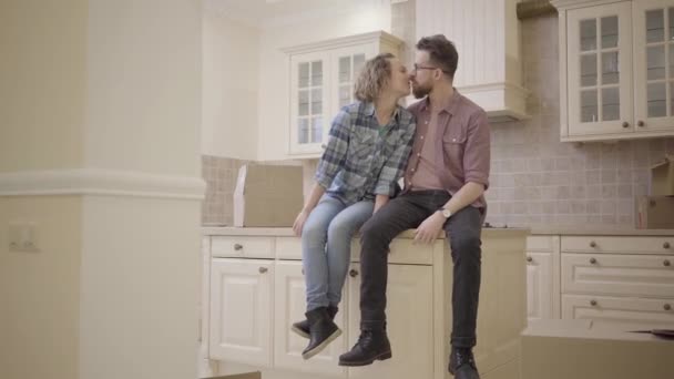 Молодая пара целуется, сидя на кухонном столе в новой квартире. Семья переезжает в новый дом. Бородатый муж в очках и кудрявая жена с светлыми волосами, одетые в повседневную одежду . — стоковое видео