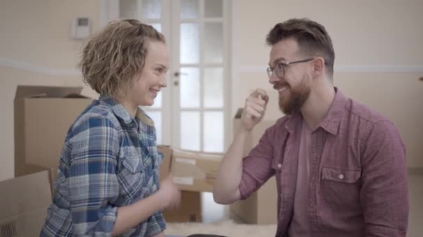 Young family sitting in light room with boxes in the background. Man showing key to woman, she stretches out hand and takes it, smiling. People huging. Married couple moves into a new home — Stock Video