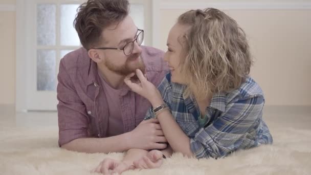 フカフカのカーペットの上に横たわる若い家族をクローズ アップ。巻き毛のプルとスクラッチのひげメガネの人の肯定的な若い女性は、彼にキスします。フレンドリーな家族時間を費やしている一緒に自宅 — ストック動画