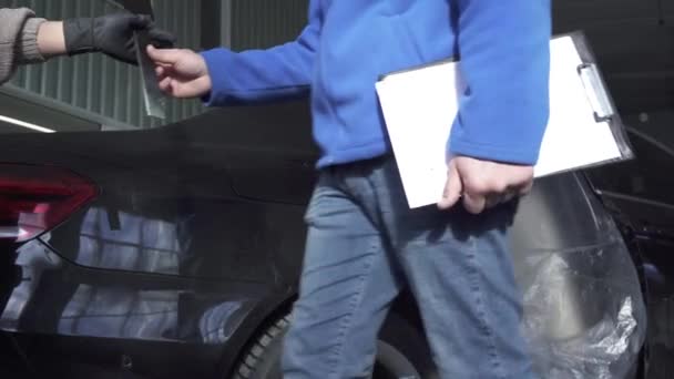 Main mechanic giving young woman an instrument with a black steel pattern for comparison with paint cars — Stock Video