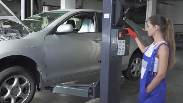 Feminino jovem automaster pressiona um botão em um mecanismo de transporte de elevação de elevação para abaixar o carro. Serviço de carro, reparação, manutenção e conceito de pessoas - mulher mecânica que trabalha na oficina . — Vídeo de Stock