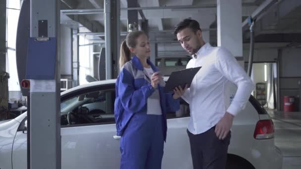 Vrouwelijke auto specialist nemen datas over auto voor de inspectie en de jonge knappe man de handtekening aanbrengend Autoservice — Stockvideo