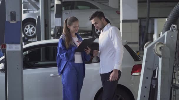 Mooi meisje auto monteur nemen datas over auto en ondertekend schudden en de hand van de jonge knappe man in autodienst nemen. Auto service, reparatie, deal en mensen concept. — Stockvideo