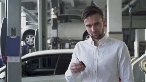 Молодий красивий чоловік в магазині автосервісу показує свої ключі від машини і посміхається на тлі своєї машини — стокове відео
