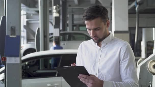 Jonge knappe man in autodienst ondertekende documenten over auto en glimlachend op de achtergrond van zijn auto — Stockvideo