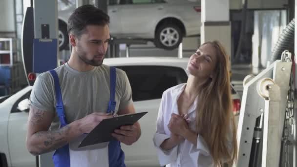 Auto-Service, Reparatur, Geschäft und Menschen-Konzept - Mechaniker und Kunde oder Autobesitzer unterzeichnen Papier in der Werkstatt. hübsche Autobesitzerin flirtet mit einem schönen Automechaniker in der Uniform. — Stockvideo