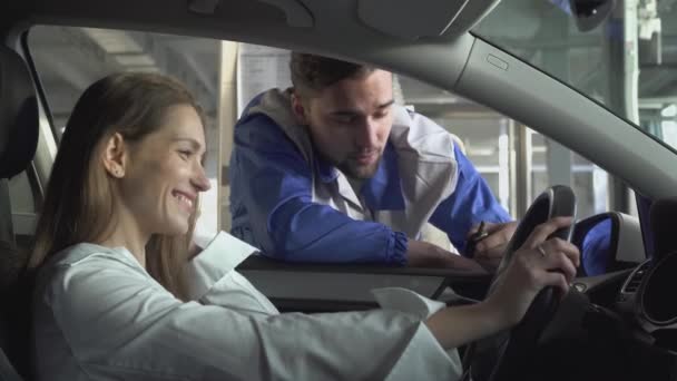 Junge Frau sitzt in einem schönen Auto, und der Autohersteller am Fenster erzählt über den technischen Teil der Benutzung des Autos nach der Reparatur — Stockvideo