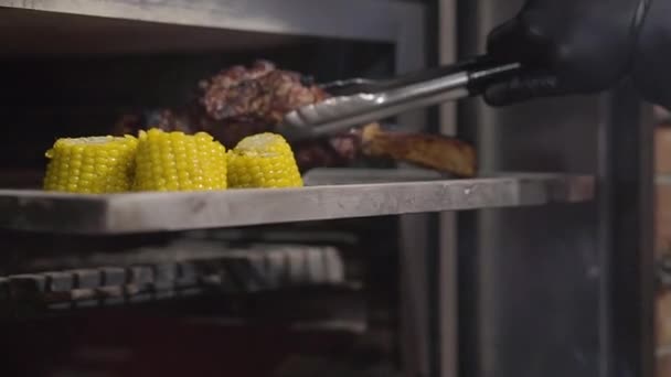 Main de cuisinier en gant noir sort juicie morceau de viande avec côtes de four à l'aide de pinces fermer. Le maïs tranché est sur une plaque de cuisson à l'intérieur du four. Au ralenti. Préparation de la nourriture dans un restaurant moderne — Video