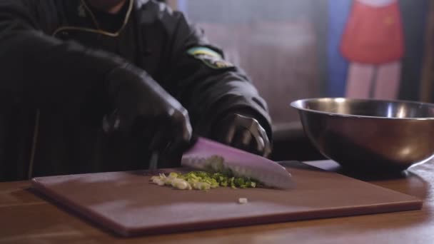 Las manos del chef en guantes de cocinero negro cortan cebolla verde sobre tabla de madera de cerca. El hombre pone cebolla cortada en un tazón grande de aluminio de pie sobre la mesa. Preparación de comida en el restaurante — Vídeo de stock