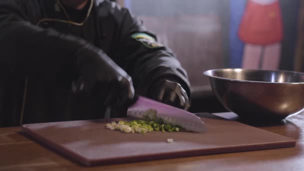 Les mains du chef en gants de cuisine noirs coupent l'oignon vert sur la planche en bois de près. L'homme met l'oignon coupé dans un grand bol en aluminium debout sur la table. Préparation de la nourriture au restaurant — Video