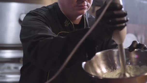 Chef en uniforme et gants de cuisine en caoutchouc noir fouettant la saucisse avec mélangeur dans un grand bol en aluminium fermer. Préparation de la nourriture dans un restaurant moderne — Video