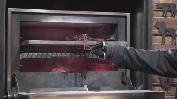 Koch in schwarzen Gummihandschuhen legt Stück Fleisch in den Ofen mit Feuer mit Metallwerkzeug in Großaufnahme. Der Koch schiebt frisches, saftiges Fleisch in den Grillofen. Zubereitung von Speisen in einem modernen Restaurant — Stockvideo