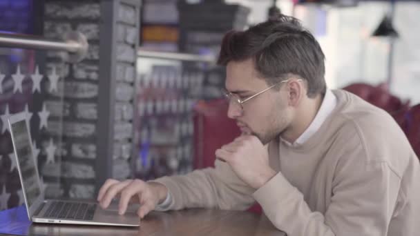 Pohledný muž sedí v kavárně, čeká na svou objednávku nebo v rozbočovači a soustředěně práci na počítači. — Stock video