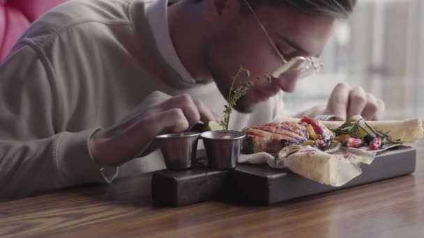 Jóképű férfi szemüveg eszik az élelmiszer, evőeszközök használata nélkül közelről. Az ügyfél grillezett hal kávézóban szolgálják fel. Magabiztos ember nyugszik a modern étterem — Stock videók