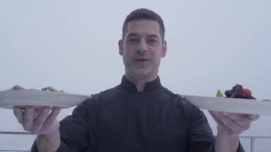 Şef siyah üniforma demostriruet iki tabak lezzetli yemekler gökyüzü açık havada bulanık bir arka plan üzerinde duran bir gülümseyen portresi sakallı.