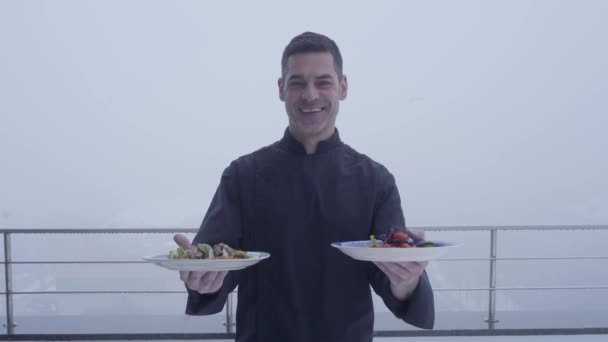 Портрет счастливого взрослого профессионального повара в черной форме с двумя тарелками на фоне дождливого туманного ландшафта — стоковое видео