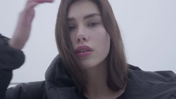 Portret van een mooie jonge meisje met donkere haren waait in de wind die permanent in een zwarte jas in mistig weer buiten — Stockvideo