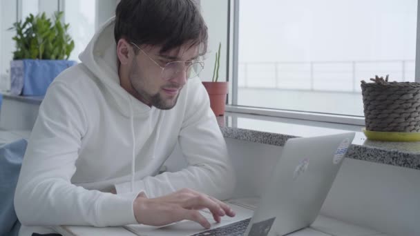 Retrato de um jovem elegante bonito barbudo cara em uma camisola leve e digitando em um laptop enquanto sentado em uma mesa à janela em um bom restaurante acolhedor ou café . — Vídeo de Stock