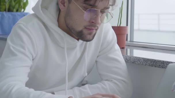 Potret pemuda berjenggot menarik dengan sweater putih dan mengetik di laptop sambil duduk di meja dekat jendela di restoran atau kafe yang nyaman . — Stok Video