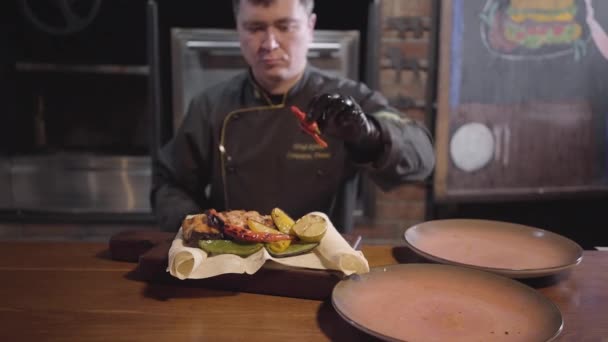Profissão chef de restaurante em uniforme preto especial serve prato de salmão peixe grelhado com legumes grelhados — Vídeo de Stock