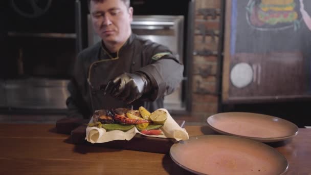 Profesión chef de restaurante en uniforme negro especial sirve plato de salmón pescado a la parrilla con verduras a la parrilla — Vídeos de Stock