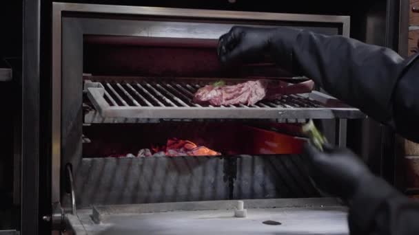 Chef-koks handen in zwarte handschoenen zetten rozemarijn op de top van de grote stuk vlees in bot in professioanal houtskool oven voor het grillen — Stockvideo