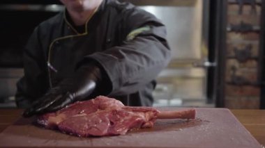 Siyah eldiven ve üniforma Restoran şefi hazırlamak çiğ et kemik, adam har vurup harman savurmak yağlı sıvı büyük parça et ile.