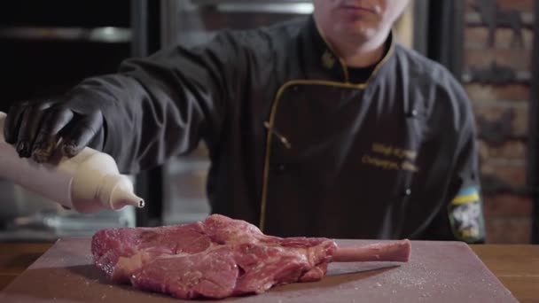 Restaurangkock i svarta handskar och uniform förbereda rått kött på benet, man pålagt stor köttbit oljig vätska. — Stockvideo