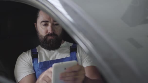 Professional mechanic sitting inside the car and verifies information on his phone. Bearded man in uniform fixing automobile in car repair station — Stock Video