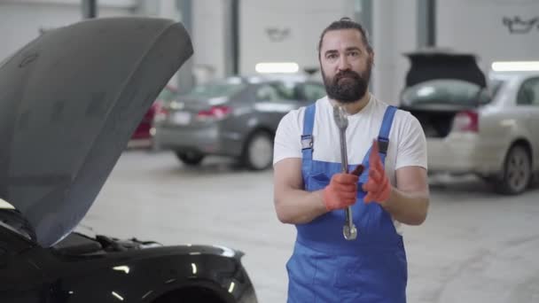 Retrato de un exitoso mecánico serio parado cerca del coche con una capucha abierta y sosteniendo una enorme llave en las manos. Barbudo hombre en uniforme de fijación de automóviles en la estación de reparación de automóviles — Vídeos de Stock