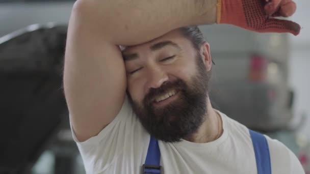 Retrato de un mecánico barbudo cansado pero sonriente, bonito y exitoso posando en guantes de trabajo y una llave inglesa en un taller de reparación de automóviles . — Vídeo de stock