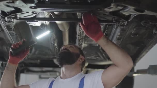 Bearded car mechanic inspecting suspension or brakes in car with lamp of lifted automobile at repair service station. Skillful man in uniform fixing car — Stock Video