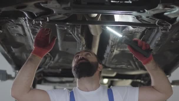 Bearded professional mechanic inspecting with lamp at repair service station. Skillful man in uniform fixing car — Stock Video