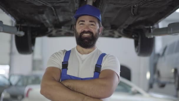 Portret przystojny Brodaty uśmiechnięty mężczyzna w postaci pracy, stojąc pod samochodu w warsztacie. Mechanik trzęsie kurz nie ręce i patrzy w kamerę. — Wideo stockowe