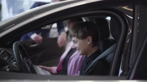 Chica bonita con el pelo corto y su madre joven sentada en un coche moderno sonriendo. Vendedor de pie cerca de la puerta abierta del automóvil. Mujer comprando vehículo para su hija — Vídeos de Stock